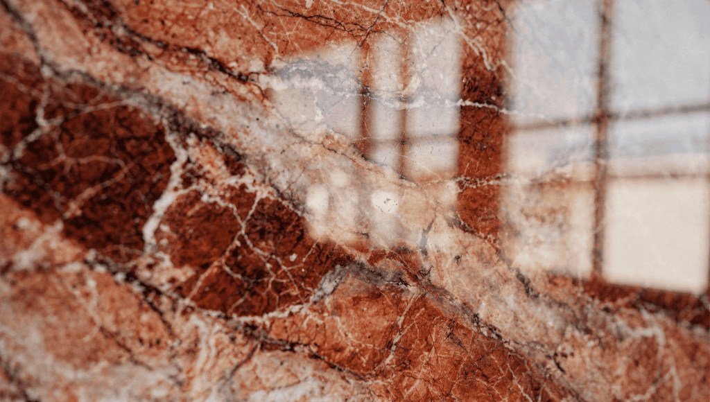 Extreme close-up of polished Rouge des Carpates marble with red terracotta tones, creamy veining, and soft window reflections.