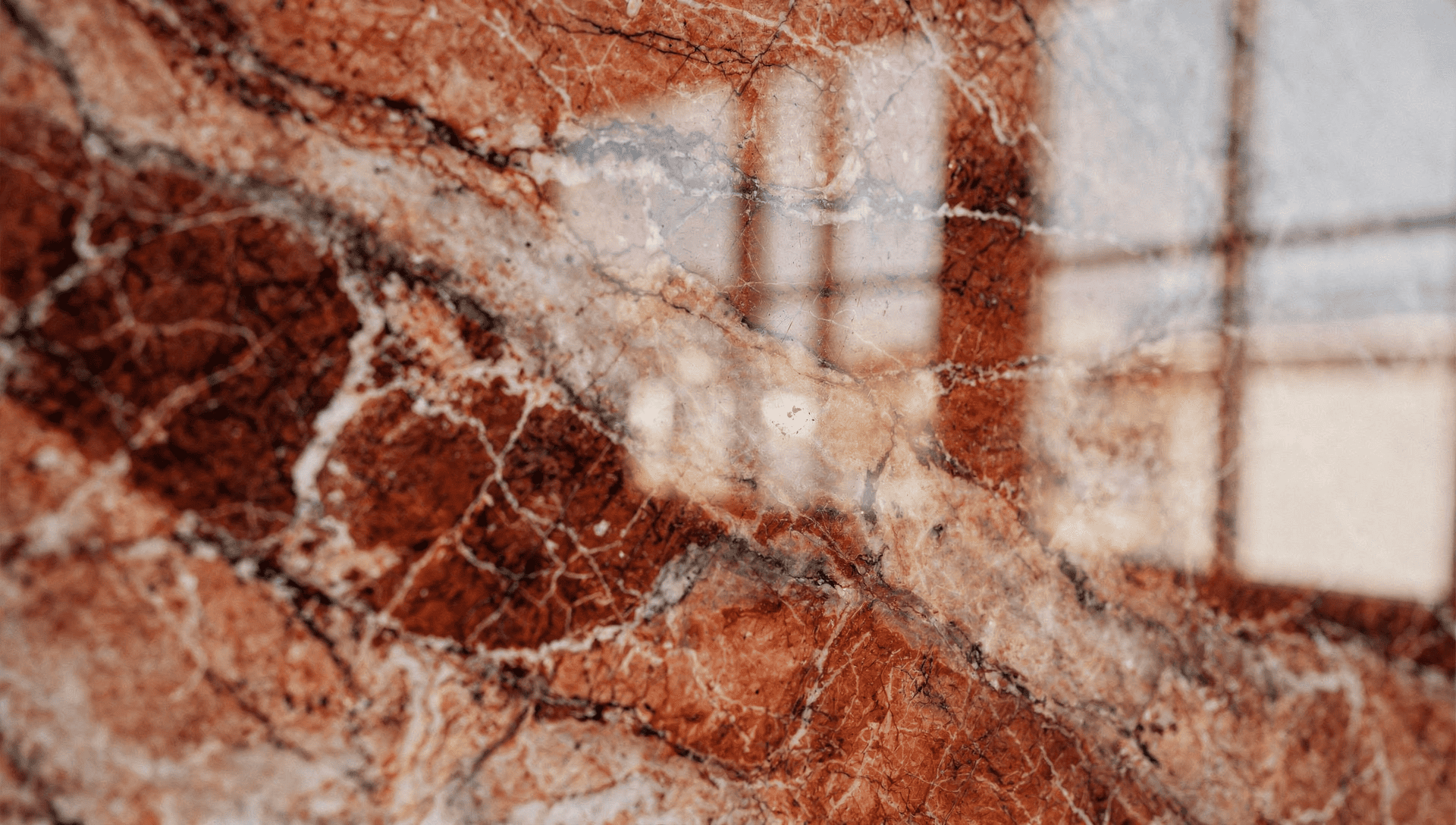 Extreme close-up of polished Rouge des Carpates marble with red terracotta tones, creamy veining, and soft window reflections.