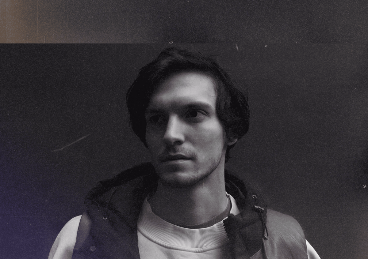 A black and white portrait of a young man Stepan Myannik with dark hair, wearing a light-colored crewneck shirt and a dark zip-up vest, set against a dark, moody background. An Artist and founder of Marmetica design