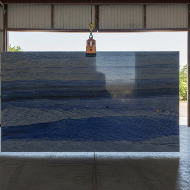 Large slab of Azul Imperial quartzite with blue banding suspended inside the hangar by a lifting clamp

